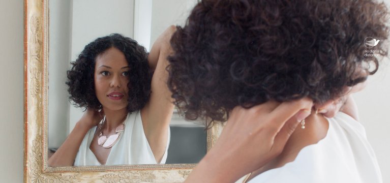 Vista trasera de una mujer con pelo rizado corto, parada frente a un espejo mirándose y con un collar