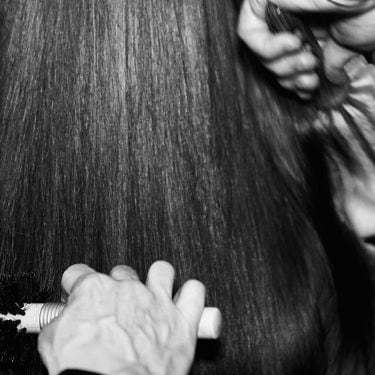 Black and white shot of the back of a woman's head with hands of a stylist with a brush and hair dryer and another stylist's hands holding a brush; both styling her hair