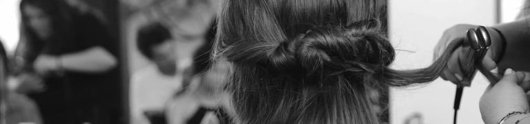 Black and white backstage close up of the back of a model's head, with her long dark hair in a loose braid, with someone straightening a section of her hair with irons